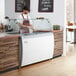 An Avantco ice cream dipping cabinet with a clear top on a counter with a man in a red apron.